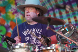 Drummer with a gray beard and wide-brim hat playing drums on a colorful tie-dye background, wearing an in-ear monitor.