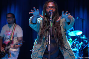 Male street performer with long dreadlocks reaching toward the camera while singing into a mic on a blue-lit stage; guitarist in background left, drummer right blurred.
