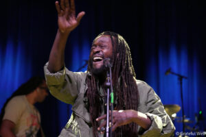 Male singer on stage with long dreadlocks, raising his right hand while singing into a microphone against a blue-lit backdrop.