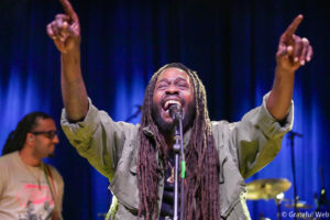 Male singer with dreadlocks on stage, arms raised, singing into a microphone against a blue curtain backdrop