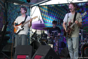 Two guitarists sing into microphones on a colorful festival stage, with a drummer in the background and tie-dye backdrop behind them.
