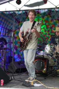 Guitarist on stage playing a dark guitar, smiling with eyes closed, colorful tie-dye backdrop behind him.