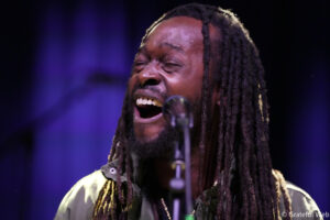 Close-up of a male singer with long dreadlocks singing into a microphone on stage, eyes closed, purple stage lighting in the background.