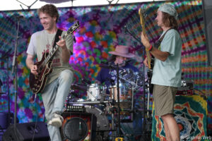 Two musicians play guitars on a colorful tie-dye stage backdrop, with a drummer in the background.