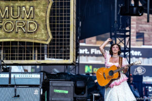 Female guitarist in a pink floral dress raises her right arm while playing an orange acoustic guitar on stage.