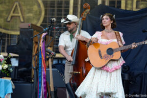 Woman in a pink floral dress playing an acoustic guitar on stage, with a bassist in a white shirt and cowboy hat nearby.