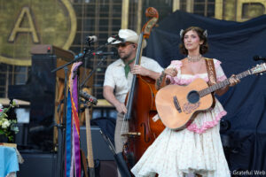 Female guitarist in a pink floral vintage dress performing on stage, with a bassist in the background.