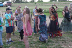 People dancing in a grassy field at an outdoor gathering, wearing colorful tie-dye shirts and flowing dresses.