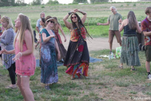 Group of adults dancing outdoors in a grassy field, wearing colorful bohemian dresses and accessories.