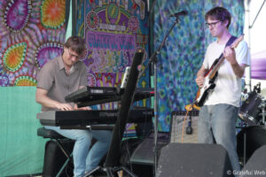 Two musicians performing on a colorful tie-dye stage: a keyboardist on the left wearing glasses, and a guitarist on the right playing an electric guitar.