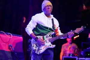 Male guitarist in a white hoodie and knit beanie, playing a sticker-covered electric guitar on stage under purple lights.