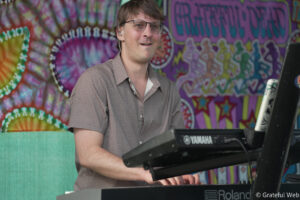 Man wearing glasses plays a Yamaha keyboard on a colorful, psychedelic stage backdrop behind him.