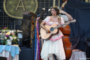 Female musician in a pink floral dress playing an acoustic guitar on stage, with a bouquet arrangement in the foreground.
