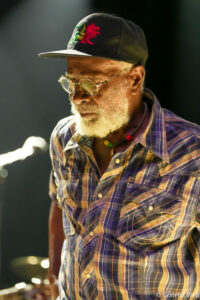 Older man with gray beard, black cap with a red dragon emblem, round glasses, and a multicolored plaid shirt on stage.