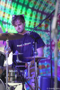 Drummer wearing a cat-ear headband playing a drum set on a colorful tie-dye backdrop