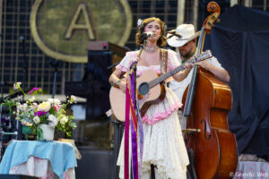 Female singer in a pink vintage dress plays an acoustic guitar and sings into a microphone on stage, with a bassist in a white hat behind her.