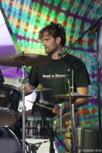 Male drummer playing a drum kit on a colorful tie-dye stage backdrop; shirt reads 'Beat a Drum' on the front.