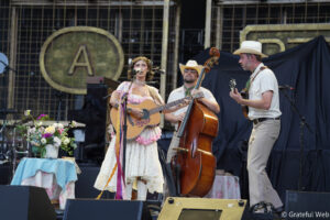 Vintage-era female singer playing acoustic guitar on stage with a banjo- or country-style band behind her; colorful flowers on a table to left.