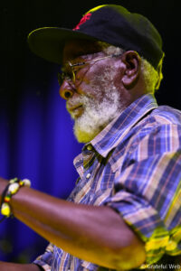 Close-up profile of an older man with a gray beard wearing glasses and a plaid shirt under stage lighting.