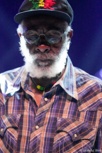 Older man with white beard wearing round glasses and a dark cap with a colorful dragon emblem, on stage in purple lighting.