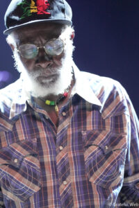 Close-up of an older man with a white beard, round glasses, and a purple plaid shirt, wearing a cap with a red dragon design.