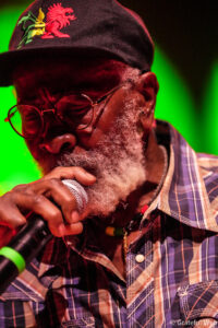 Older man with a gray beard and glasses singing into a handheld microphone on stage, wearing a plaid shirt and a cap with a dragon embroidery.