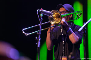 Musician wearing a cap and glasses playing a trombone on stage with green lights in the background.