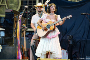 Female musician in a pink floral vintage dress playing an acoustic guitar on stage, with a guitarist in the background and multiple microphones around.