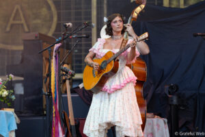 Female musician in a pink floral dress playing an acoustic guitar on stage during a live performance.