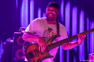Smiling bass guitarist performing on a stage bathed in purple light.