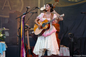 Female musician in a pink, lace dress plays an acoustic guitar on stage, with microphones nearby and stage backdrop visible.