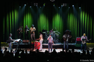 Band performs on a dimly lit stage with green curtains and spotlights, musicians playing keyboards, saxophones, drums, guitar, and bass.