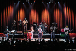 A live band performs on a lit stage with red curtains, players playing keyboards, saxophones, drums, guitar, and a vocalist front and center while an audience watches from the darkened foreground.
