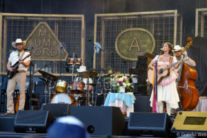 Female singer in a vintage floral dress sings and plays acoustic guitar on stage, with a male guitarist and drummer behind her.