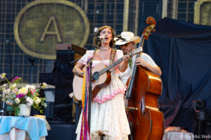 Female singer in a floral vintage dress playing an acoustic guitar and singing into a mic on stage, with a double bass player in the background and flowers on a table to the left.
