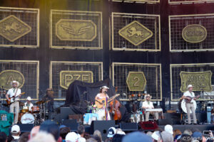 Outdoor concert stage with a country band performing; musicians play guitars and drums, wearing cowboy hats, in front of large decorative metal panels with crest-like emblems. A cheering crowd is visible in the foreground.