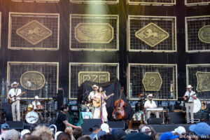 Live outdoor concert with a band performing on a large stage, musicians in western attire; center stage features a female singer/guitarist with flowers, others on guitars, drums, and keyboard against decorative panels with logos behind them.