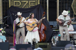 Female guitarist in a pink floral dress sings on stage while backing musicians play guitar and bass behind her.