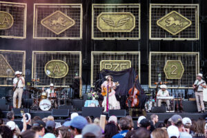 Outdoor concert stage with a band performing; a female guitarist/vocalist center stage, drummer and other musicians to her sides, audience in the foreground; gold decorative panels with heraldic emblems backdrop.