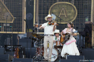 Male violinist in a white shirt and cowboy hat performs on stage beside a female guitarist in a pink floral dress; drums and microphones around.