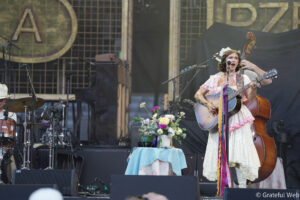Female singer and guitarist in a vintage pink dress performing on an outdoor stage, with flowers on a table in front.