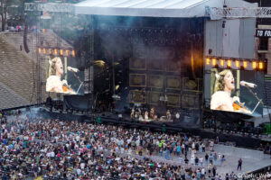 Outdoor concert in a stadium with a large stage, two giant video screens, and a cheering crowd.
