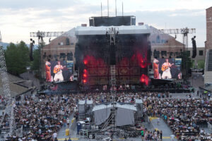 Outdoor concert at a large stadium with a grand stage, oversized video screens, and a cheering audience in front of the stage.