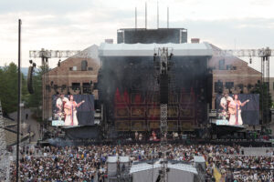 Large outdoor concert stage with tall rigging, two big LED screens on either side showing performers, and a dense audience in front.