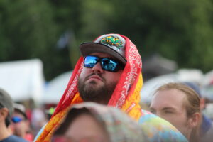Bearded man in dark cap and blue mirrored sunglasses wrapped in a bright red and orange patterned scarf at a crowded outdoor event.