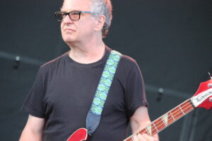 Male musician wearing a black T-shirt and glasses playing a red bass guitar on stage, with a patterned green strap visible against a dark background.