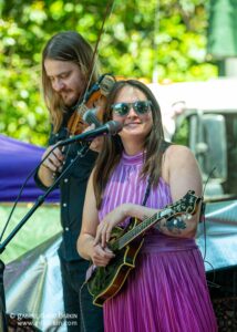 Female musician singing into microphone while playing an acoustic guitar, wearing a purple pleated dress and sunglasses, outdoors on stage.