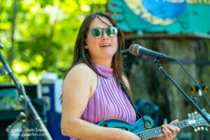 Female guitarist and singer performing outdoors, wearing a pink pleated top and mirrored sunglasses while playing a blue guitar.