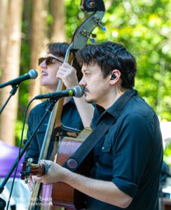 Musician singing into a microphone while playing an acoustic guitar outdoors, with another guitarist and a bass visible in the background.