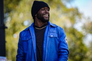 Man in a bright blue jacket and black beanie smiles outdoors, autumnal trees blurred in the background.